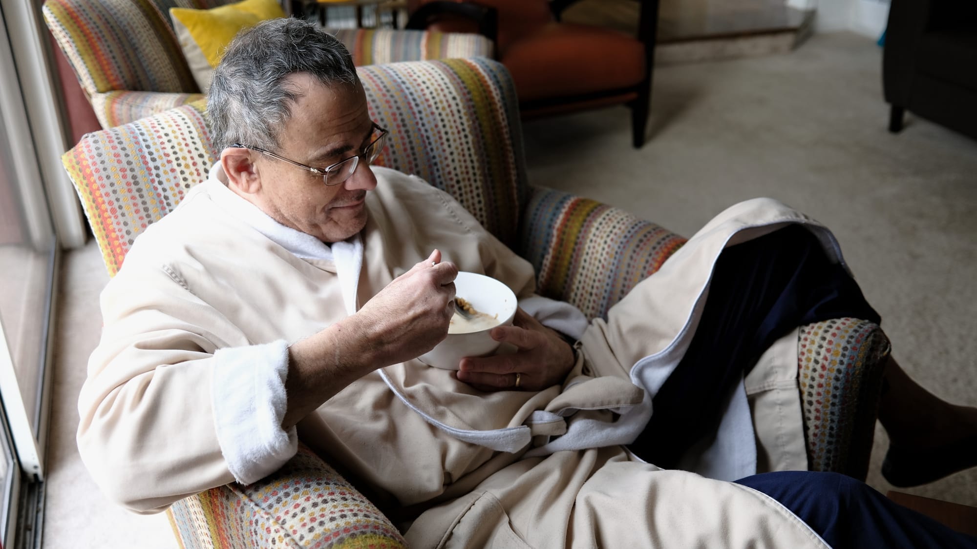 A man sits in a chair with his leg over the arm while he eats cereal.