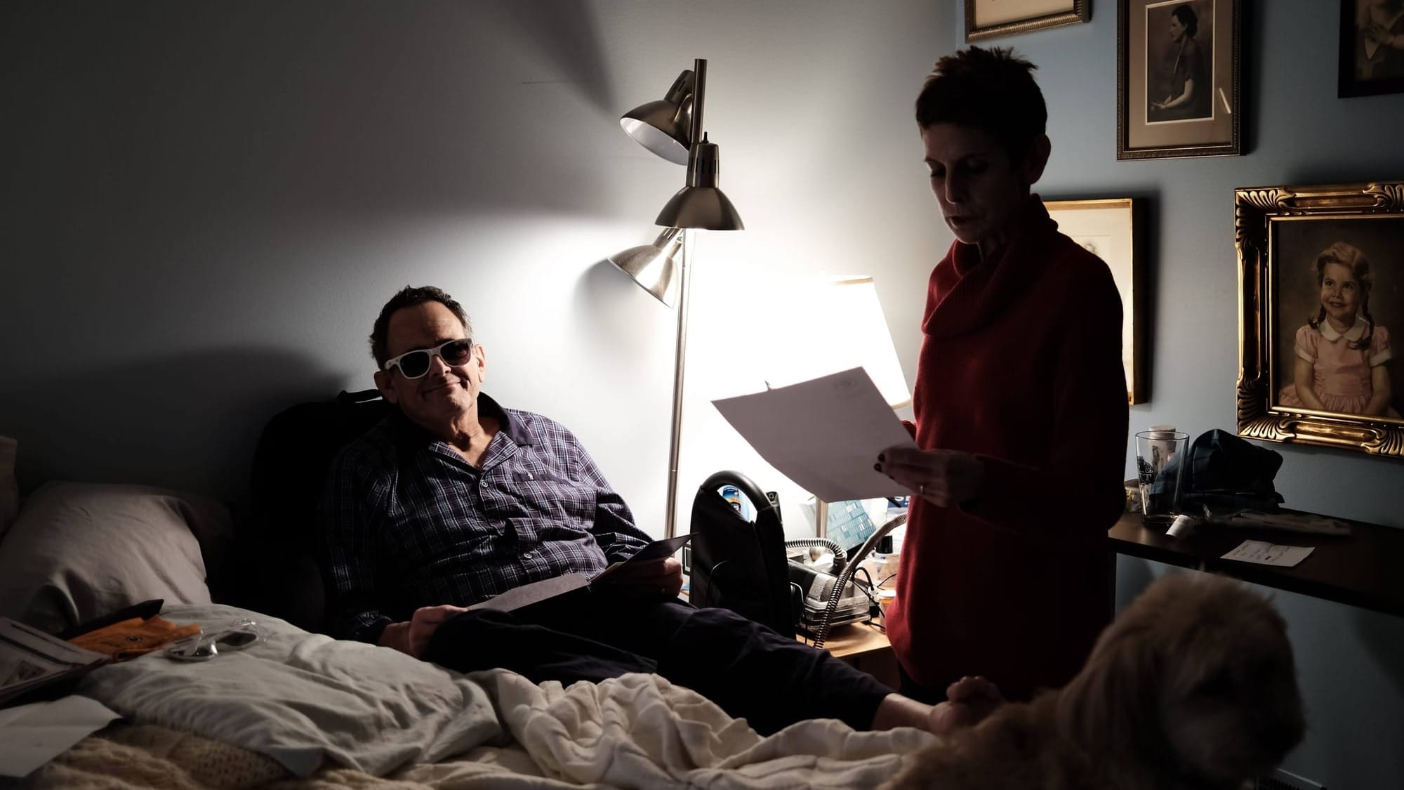 A man wears sunglasses indoors while a woman stands holding a sheet of paper.