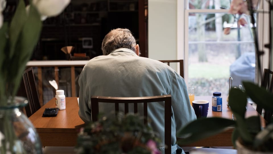 A man sits at a kitchen table, facing away from the camera.